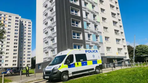 Blue and yellow check police, and a police officer, outside a large building, under blue skies.