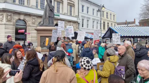 Richard Roberts A large crowd of people outside some white-fronted buildings with a number holding up white placcards
