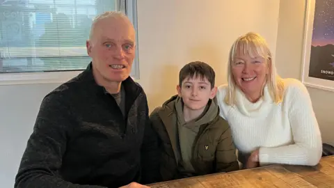 Hywel and Mandy Jones with George, 11, sat a wooden cafe table. Hywel and George are wearing dark jumpers and jackets, while Mandy is wearing a white wool jumper. They are all smiling at the camera.