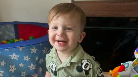 Hudson smiles at the camera with a ball pit behind him. He is wearing a light green shirt with micky mouse faces all over it. Hudson has brown-ginger hair.