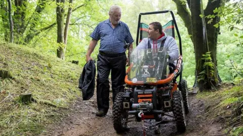 Submitted/George Pas George Pas wearing a grey hoodie and driving through the forest on his orange Overlander off-road wheelchair. Walking beside him is his father, wearing black trousers and a blue and white checked shirt. They are chatting and looking at the each other with affection.