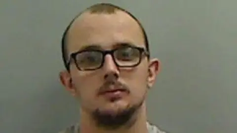 Mugshot of Cooper. He has a blading head of dark hair, dark goatee and moustache with black glasses