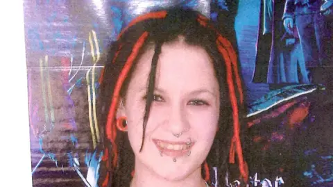 A woman with red and black dreadlocks and several lip and nose piercings smiles.