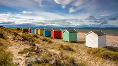 Getty Images A row of colourful beach huts at Findhorn. The colours of the little, wooden structures include white, green, red, blue and yellow. Below the huts is the beach and sea beyond. It is a sunny day with clouds int he sky.