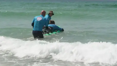 Surfing England Cam Treble is seen on a board being supported by two men wearing blue T-shirts in the waves 