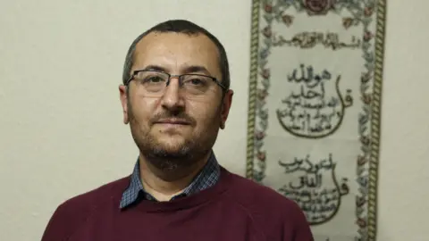 Kateryna Pavlyuk A man with dark cropped hair and a short beard stands in front of an Arabic wall hanging on a pale, textured wall. He wears rectangular glasses and a burgundy jumper, looking to camera with a serious expression.