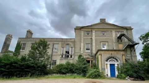 Steve Huntley/BBC The former Moreton Hall School a Georgian-style manor house. On the right is a three storey building and extending on the left is a two storey wing. It has smashed windows and appears derelict. Trees and shrubs have grown up on either side of the portico style front door, which is on the right. In front of that is gravel and resting on the gravel, on the right is a pile of burnt out debris, including a blackened sofa frame.