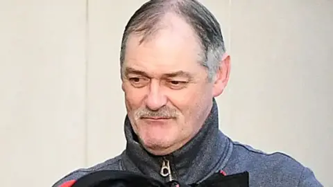 Daniel McClean. He has grey hair and a moustache, brown eyes and wearing a grey zip up jacket. 