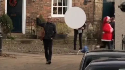 BBC Mr Sutherland walks down a path with a brown bag slung over his shoulder. He is wearing sunglasses and is dressed in dark clothes, with one hand in his pocket and the other raised in a thumbs-up. A microphone can be seen in the right corner.