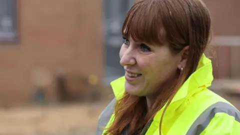 Angela Rayner, in a yellow high-vis jacket, looks to the left of the shot and is smiling. She has long brown hair and has a gold-hoop earring.