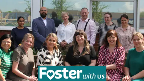 The Foster With Us team. The staff have are lined up in two rows, one behind the other, behind a teal sign reading foster with us.