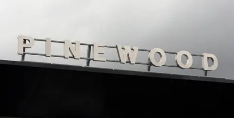 PA Media The front gate of Pinewood studios in Iver Heath. The word Pinewood has been fastened to a metal frame. They stand out against a grey sky. 