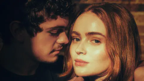 Helen Murray Noah Jupe has brown, curly hair and he is looking at Sadie Sink, who has red hair and is looking at the camera.  