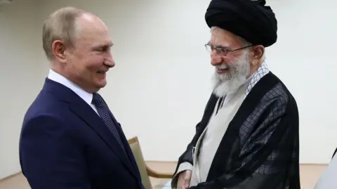 Putin on the left shakes the hand of Ayatollah Khamenei. Both men are smiling. Putin is wearing a navy suit while Khamenei is wearing traditional Islamic clerical robes and a black turban, along with glasses.