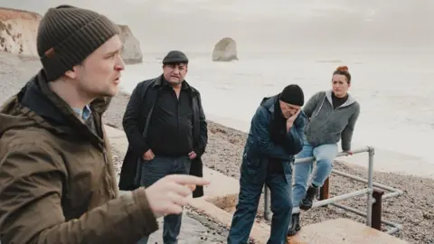 Charlie Price Four of the actors photographed in the bay on some steps. The beach is made of stones, and coastal cliffs are in the background.