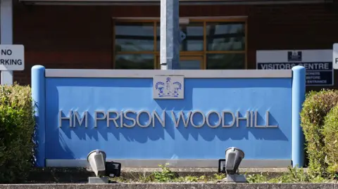 Blue sign with grey writing reading HM Prison Woodhill. There are lights at the bottom of the sign pointing upwards and hedges either side. 