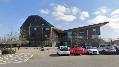 Google A modern-looking, dark coloured building with a sloping roof and cars parked outside. 