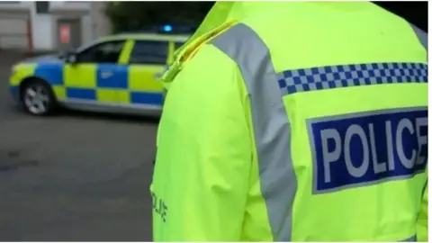 View of the back and shoulders of a police officer wearing a high visibility jacket. To the left, and slightly out of focus, is a police vehicle.