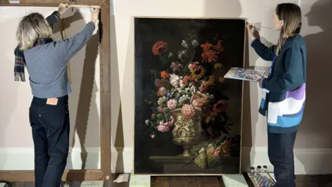 Critchlow & Kukkonen Ltd Two women stand in front of an artwork depicting a large vase of pink, orange and red flowers, against a black background. One holds a paint tray and a paintbrush, while the other holds a measuring tape.