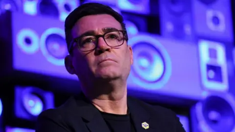 Andy Burnham looks serious while standing in front of a blue and purple background.