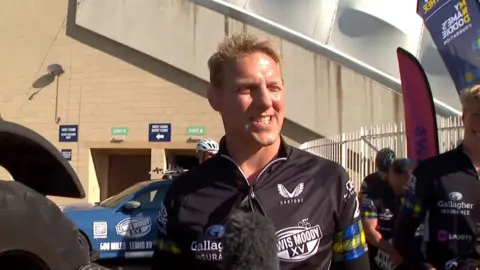 Lewis Moody, a man with short blonde hair, stands looking into the sun, smiling, dressed in cycling gear