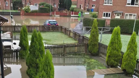 Backyards filled with standing water on a residential estate. Flooding can be seen in gardens and on a nearby street.