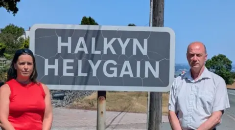 A woman in a red top and brown hair and a balding man in a pale blue shirt. They're stood outside a village in front of a sign that says 'Halkyn Helygain' . 
