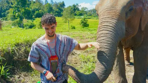 Calum Macdonald pets an elephant as it touches his striped patterned shirt with its trunk. Behind them are green paddy fields and tropical woods beyond that. He has short black curly hair.