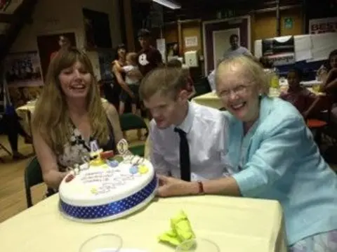 Rosalie Lander Charlie, his sister and mum, sit around a decorated birthday cake with candles, while other guests are visible in the background