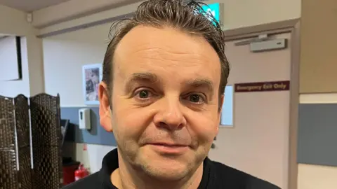 Man looking directly at the camera with brown hair, brown eyes and a soft smile. 
He is wearing a black turtleneck jumper and standing in a dressing room. 