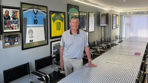 Ray Parr is leaning against a long table in the Striker's Lounge. The table is covered in a blue and white chequered table cloth and on the walls is football memorabilia including signed shirts and photographs.