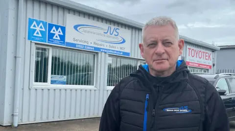 A man with short grey hair, and wearing a dark-blue anorak with blue "JVS" logo, stands in front of a garage - a squat, metal-clad building with rectangular windows and a sign reading "JVS, Japanese Vehicle Specialists, Serving, Repairs, Sales, MOT".