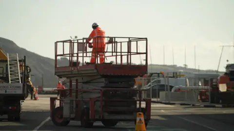 A mobile lift operator in an orange outfit on a dockside.