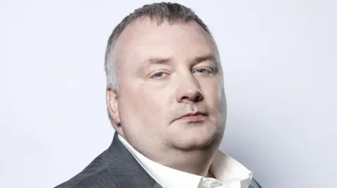 BBC A man with short hair is turned to the side looking into the camera. He is wearing a white shirt and grey blazer.