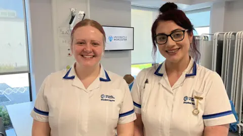 BBC Two women in a mainly white uniform including the words University of Worcester Student Nurse. Windows and walls are behind them and there is a screen on the wall featuring the words University of Worcester.