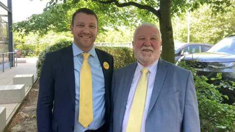 Emma Howgego/BBC Henry and John Batchelor stare at the camera. They both wear matching yellow ties. Henry, on the left, wears a navy suit with a light-blue shirt and a Liberal Democrat badge. John, on the right, wears a light-grey suit and white shirt.
