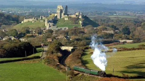Andrew PM Wright Aerial view of a steam locomotive and four green carriages winding its way through the valley beneath the hilltop ruins of Corfe Castle.