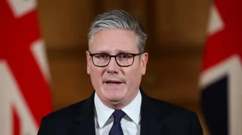 Keir Starmer. He has short grey-ish hair, wearing glasses, white shirt and dark blazer and tie. Two British flags are in the background.