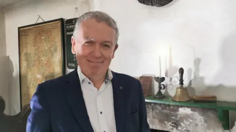 A man stands in front of a fireplace, upon which sits a bell, a a candle in a candlestick holder and a piece of wood that looks like a blackboard wiper. The man wears a navy blazer over and white shirt and is smiling. Over his left shoulder is a browned map of Ireland.
