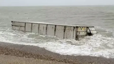 A white shipping container in the surf off a stoney beach