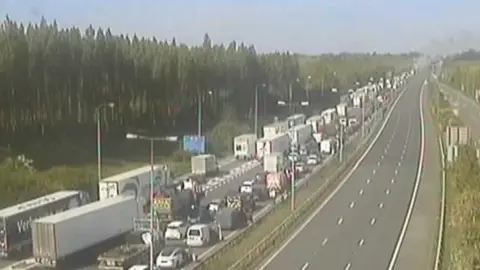 National Highways National Highways CCTV footage showing traffic building up toward the fire, with cars and lorries stuck in standstill while emergency services work on ensuring people's safety. The sky is clear and blue with smoke rising up on the horizon in the background.
