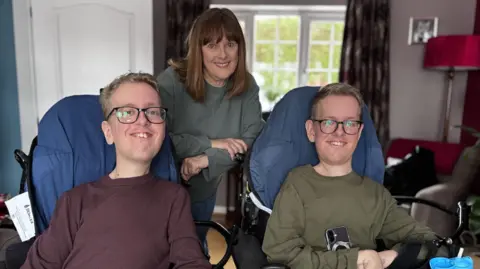 Identical twins Sam and Alex Bolton pictured sat in their wheelchairs in their living room smiling, with their mum stood behind them leaning on the wheelchair. 