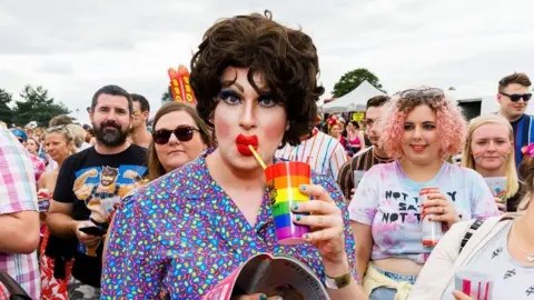 Martin Parr/Magnum Photos A drag queen wearing a colourful blouse, brown curly wig and bright, red lipstick at Bristol Pride. She is holding a rainbow coloured cup and sipping the yellow straw. There are other people around her wearing colourful clothes.