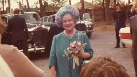 Cynthia Spencer Hospital Queen Elizabeth, the Queen Mother, standing outside, wearing a flowered hat, holding a bouquet for flowers, and wearing a blue suit with a broach. She is standing by cars, police are to the right and other people to the left and infront. 