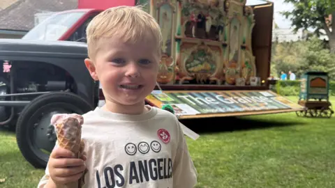 Louise Parker/BBC A three-year-old child with an ice cream. In the background is a Dutch organ on a trailer.