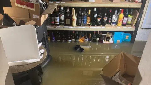 Lee Maloney A flooded cellar full of bottles of spirits and cardboard boxes. Murkey greeny-brown water is on the floor.