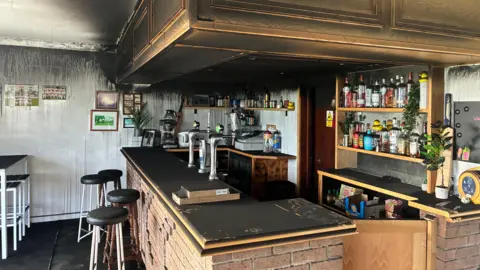 The interior of a clubhouse bar covered in black soot. There is a bar and several stools.