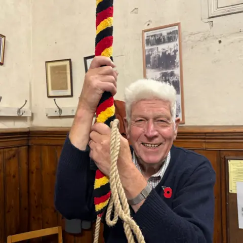 Andy Vivian Jack is smiling to camera in the bell tower, holding onto a rope which is black, red and yellow-striped. He has white hair and is wearing a blue jumper with a poppy attached. There is a black and white photo behind him showing old pictures of bell-ringers