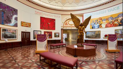 David Rowan/Birmingham Museums Trust A circular room with a golden statue of an angel in the centre. The angel is surrounded by red benches and has two floor-standing plaques on either side of him. There are numerous pieces of art on the walls and at the top of the walls on either side of the image are the words "A fresh take"