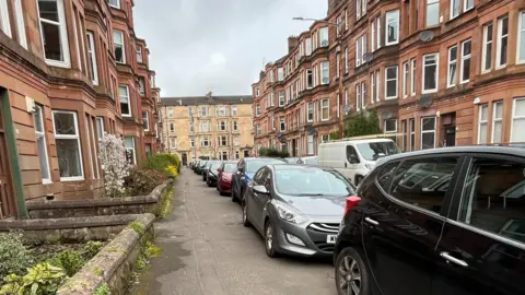 A narrow residential street lined with parked cars on both sides, bordered by tall red‑sandstone tenement buildings. The pavement is wet, the sky is overcast, and plants and shrubs appear along the left side near the building entrances.